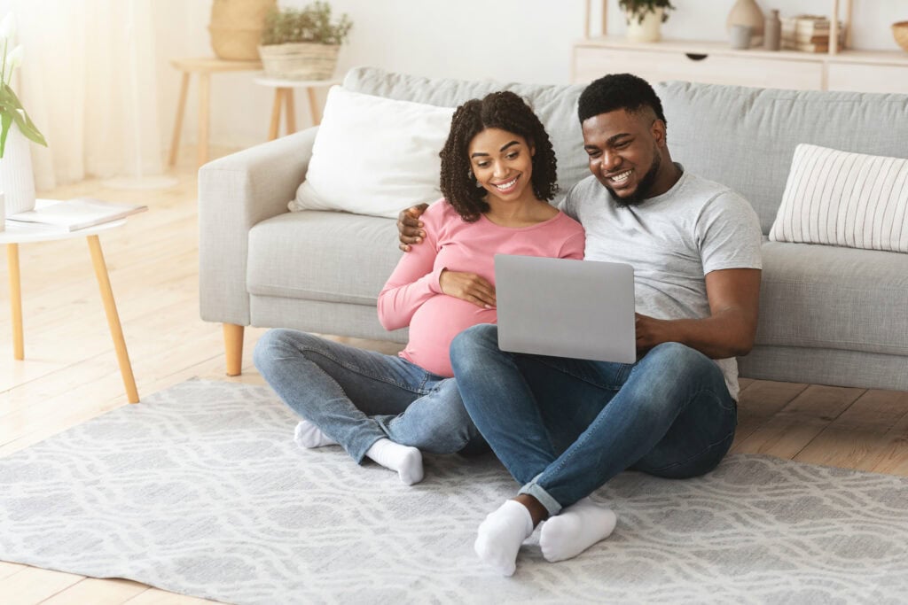 Cheerful expecting black family spending time together at home, hugging and using laptop, copy space